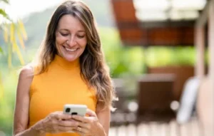 Mujer sonriendo mientras mira su celular, disfrutando del momento tras activar tu red gratis en un entorno al aire libre.