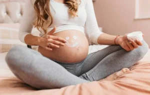 Mujer embarazada sentada en la cama, aplicando crema hidratante en su vientre con una carita sonriente dibujada, expresando ternura y conexión con tu bebé.