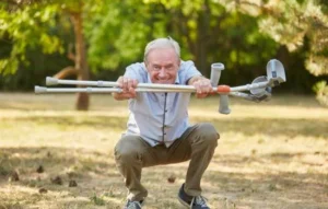 Hombre mayor sonriendo mientras se agacha y levanta sus muletas en señal de superación, rodeado de naturaleza, demostrando que es posible aumentar su vitalidad.
